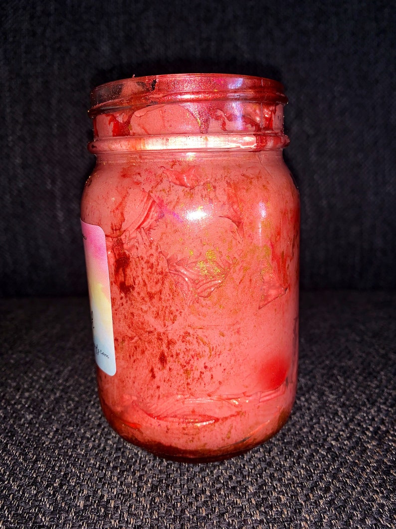 May include: A glass jar with a red, pink, and gold swirl design. The jar is filled with a red, pink, and gold substance. The jar has a white label with the text "The Good Witch" on it.