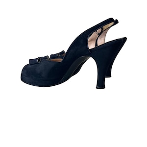May include: A black suede slingback high heel shoe with a rounded toe and a small platform sole. The shoe features a decorative bow detail on the front and a thin heel. The interior lining is a light beige color, visible at the heel and toe area.
