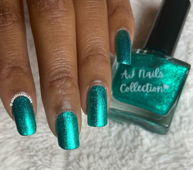 May include: A teal green nail polish bottle with a clear glass bottle and a black cap. The bottle has a label that says "AJ Nails Collection" with a heart symbol. The image shows a hand with four fingernails painted with the teal green nail polish.