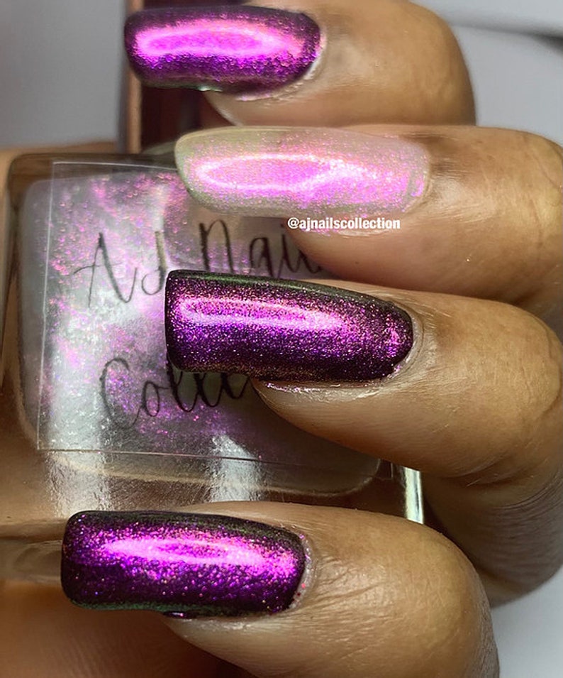 Puede incluir: Primer plano de una mano con cuatro u&ntilde;as pintadas con un esmalte de u&ntilde;as iridiscente de color morado oscuro. El esmalte de u&ntilde;as tiene un acabado brillante y refleja diferentes colores a la luz. La mano sostiene una peque&ntilde;a botella de esmalte de u&ntilde;as con el texto "AJ Nails Collection" en la etiqueta.