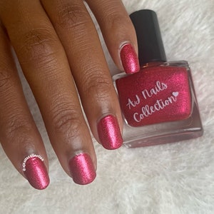 Può includere: Una mano con unghie curate dipinte con uno smalto rosso scintillante. Una boccetta di smalto con tappo nero e la scritta "AJ Nails Collection" è accanto alla mano.