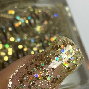 May include: Close-up of a fingernail painted with gold glitter nail varnish. The varnish has a clear base with gold and iridescent hexagonal glitter. The nail varnish bottle is visible in the background. The text "@ajnailscollection" is on the nail.