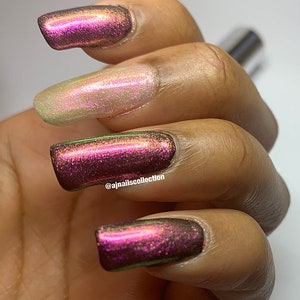 Puede incluir: Primer plano de una mano con cuatro u&ntilde;as pintadas con un esmalte de u&ntilde;as de color p&uacute;rpura oscuro iridiscente. El esmalte de u&ntilde;as tiene un brillo sutil y un tono verde. La mano est&aacute; apoyada sobre una superficie blanca. El nombre de usuario de Instagram @ajnailscollection es visible en el dedo anular.