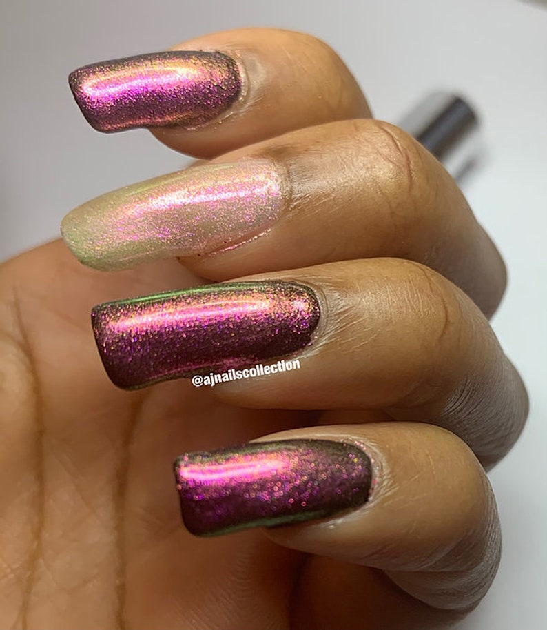 Puede incluir: Primer plano de una mano con cuatro u&ntilde;as pintadas con un esmalte de u&ntilde;as de color p&uacute;rpura oscuro iridiscente. El esmalte de u&ntilde;as tiene un brillo sutil y un tono verde. La mano est&aacute; apoyada sobre una superficie blanca. El nombre de usuario de Instagram @ajnailscollection es visible en el dedo anular.