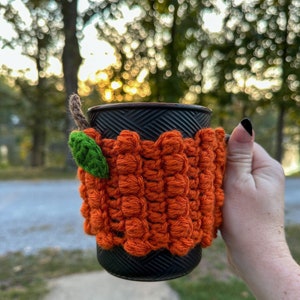 May include: A black mug with a crocheted orange pumpkin-shaped cosy. The cosy has a green leaf detail.