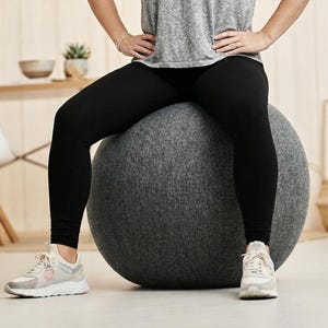 Op de afbeelding: Een grijze fitnessbal is het middelpunt van de afbeelding. De persoon draagt een grijs t-shirt, zwarte leggings en witte sneakers. De achtergrond omvat een witte stoel, een kleine tafel en een ingelijste foto.
