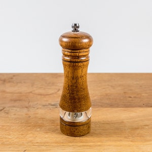 K&ouml;nnte beinhalten: Eine Pfefferm&uuml;hle aus Holz mit einem transparenten Acrylfenster und einem silbernen Band. Die Pfefferm&uuml;hle besteht aus hellbraunem Holz und hat einen silbernen Deckel. Sie steht auf einer Holzoberfl&auml;che.