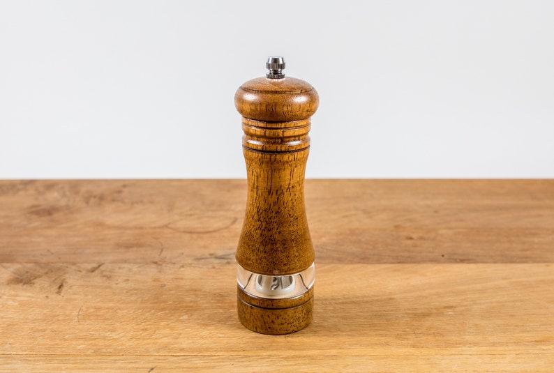 K&ouml;nnte beinhalten: Eine Pfefferm&uuml;hle aus Holz mit einem transparenten Acrylfenster und einem silbernen Band. Die Pfefferm&uuml;hle besteht aus hellbraunem Holz und hat einen silbernen Deckel. Sie steht auf einer Holzoberfl&auml;che.