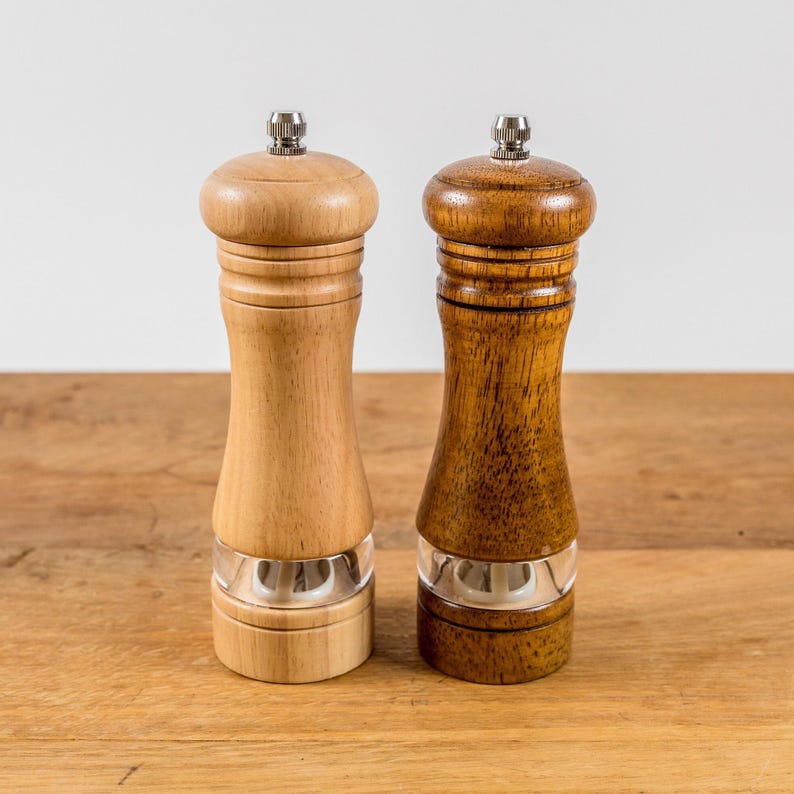 K&ouml;nnte beinhalten: Zwei Salz- und Pfefferm&uuml;hlen aus Holz. Eine ist hell, die andere dunkelbraun. Beide haben einen transparenten Acrylabschnitt am Boden und eine silberfarbene Oberseite. Sie werden auf einer Holzoberfl&auml;che pr&auml;sentiert.