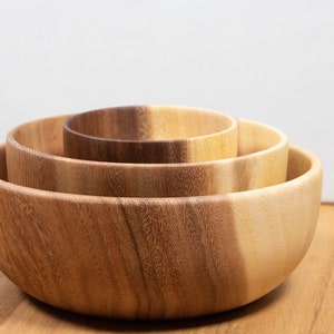 May include: Three wooden bowls of varying sizes nested together. The bowls are made of light brown wood with a natural grain pattern. The bowls are arranged in a stack, with the largest bowl at the bottom and the smallest bowl at the top.