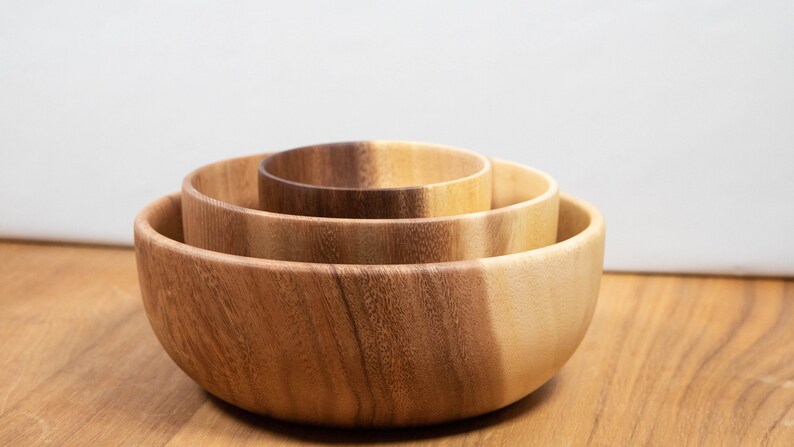May include: Three wooden bowls of varying sizes nested together. The bowls are made of light brown wood with a natural grain pattern. The bowls are arranged in a stack, with the largest bowl at the bottom and the smallest bowl at the top.