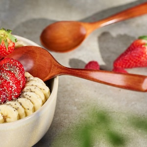 May include: A beige bowl filled with sliced strawberries, bananas, raspberries, and chia seeds. A fig and a whole strawberry are also visible. Two wooden spoons rest on the surface, suggesting a healthy breakfast or snack.