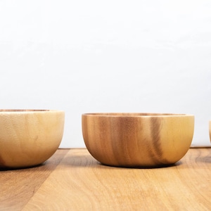 May include: Three wooden bowls of varying sizes are arranged on a wooden surface. The bowls are made of light and dark brown wood, with a smooth, curved design. The bowls are empty and ready for use.