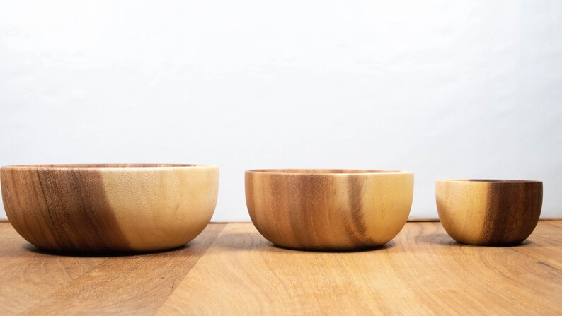May include: Three wooden bowls of varying sizes are arranged on a wooden surface. The bowls are made of light and dark brown wood, with a smooth, curved design. The bowls are empty and ready for use.