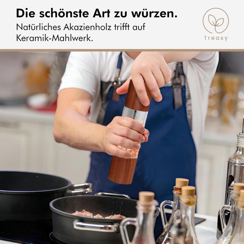 May include: A wooden salt or pepper grinder with a clear glass body is being used over a pan. The grinder has a dark brown wooden top and bottom. The text "Die sch&ouml;nste Art zu w&uuml;rzen" is visible.