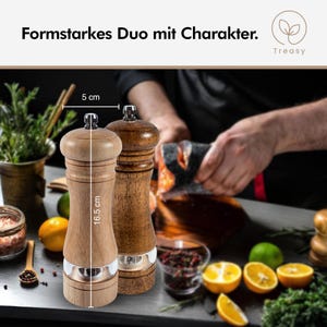 K&ouml;nnte beinhalten: Zwei h&ouml;lzerne Pfefferm&uuml;hlen, eine helle und eine dunkle, mit silbernen Akzenten. Die gr&ouml;&szlig;ere M&uuml;hle ist 16,5 cm hoch und die Basis 5 cm breit. Der Text "Formstarkes Duo mit Charakter" steht oben.