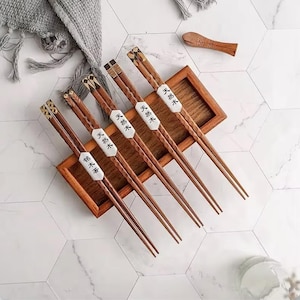 May include: A set of wooden chopsticks with decorative ends and white labels with Asian characters. The chopsticks are displayed in a wooden tray with a small wooden spoon. The background is a white tile surface.