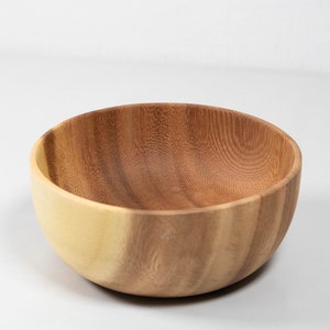 May include: A wooden bowl with a natural wood grain finish. The bowl has a light-colored exterior and a darker interior, showcasing the natural variations in the wood. The bowl is round and appears to be handcrafted.