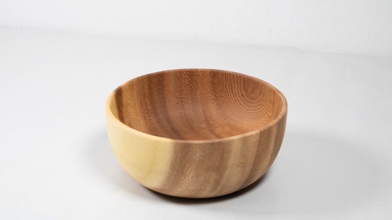 May include: A wooden bowl with a natural wood grain finish. The bowl has a light-colored exterior and a darker interior, showcasing the natural variations in the wood. The bowl is round and appears to be handcrafted.