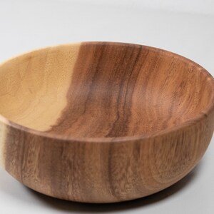 May include: A wooden bowl with a natural finish. The bowl is round with a smooth interior and exterior. The wood grain is visible, with variations in color from light to dark brown.