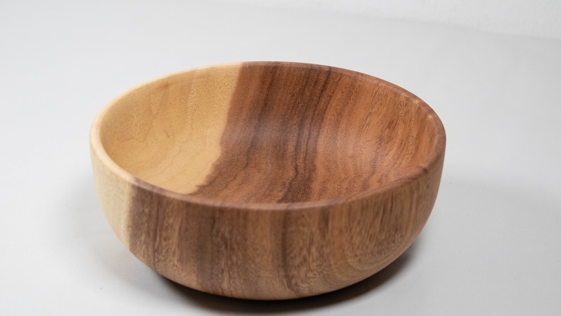 May include: A wooden bowl with a natural finish. The bowl is round with a smooth interior and exterior. The wood grain is visible, with variations in color from light to dark brown.