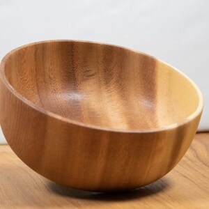 May include: A wooden bowl with a slanted rim, crafted from light brown wood. The bowl is empty and sits on a wooden surface, showcasing the natural grain and color variations of the material. The background is a plain white.