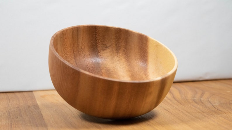 May include: A wooden bowl with a slanted rim, crafted from light brown wood. The bowl is empty and sits on a wooden surface, showcasing the natural grain and color variations of the material. The background is a plain white.
