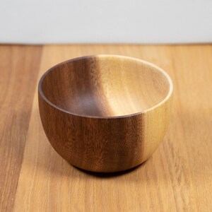 May include: A small, round wooden bowl with a natural finish. The bowl is made of a light-colored wood and has a smooth, polished surface. It sits on a wooden surface.