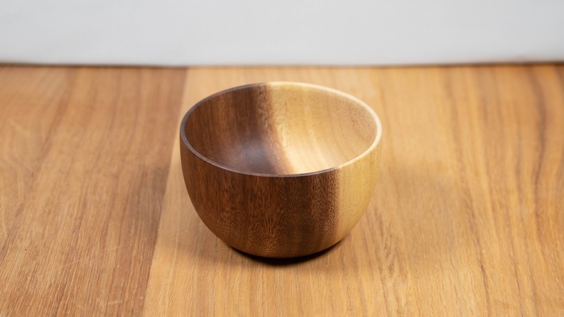May include: A small, round wooden bowl with a natural finish. The bowl is made of a light-colored wood and has a smooth, polished surface. It sits on a wooden surface.