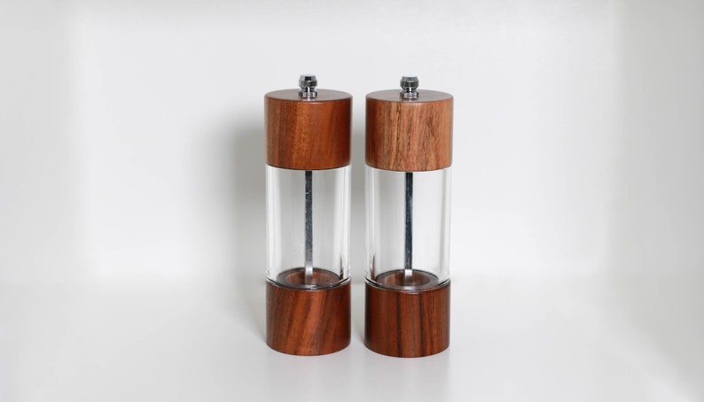 May include: Two wooden salt and pepper grinders with clear acrylic bodies. The grinders have a dark wood finish on the top and bottom, with a silver metal mechanism. The background is white.