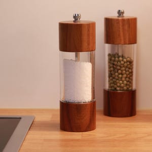 Puede incluir: Dos molinillos de sal y pimienta de madera con cuerpos de acrílico transparente. Un molinillo contiene sal blanca, el otro granos de pimienta verde. Los molinillos tienen un diseño clásico con tapa y base de madera y tapa de metal plateado.