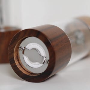 May include: Close-up of a wooden salt or pepper grinder with a clear glass body. The grinder has a dark brown wooden top and base, with a silver grinding mechanism visible. Another grinder is blurred in the background.