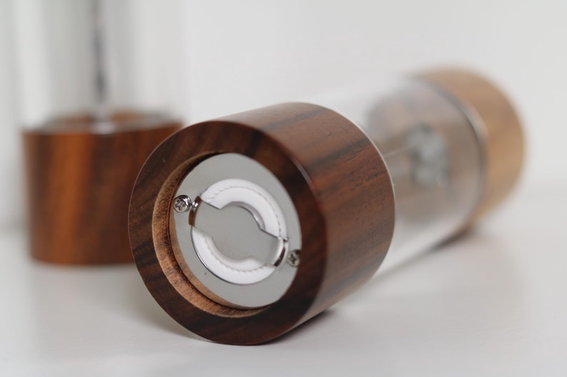 May include: Close-up of a wooden salt or pepper grinder with a clear glass body. The grinder has a dark brown wooden top and base, with a silver grinding mechanism visible. Another grinder is blurred in the background.
