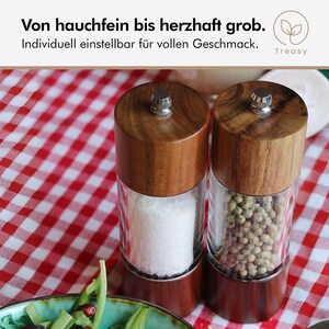 May include: Two wooden salt and pepper grinders with clear glass bodies, one filled with white salt and the other with green peppercorns. The grinders sit on a red and white checkered tablecloth. The text at the top says, "Von hauchfein bis herzhaft grob."