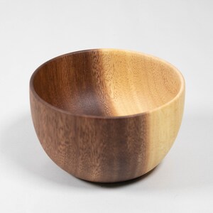 May include: A wooden bowl with a natural wood grain finish. The bowl has a rounded shape and a smooth interior. The color is a mix of light and dark brown tones. This bowl is suitable for serving food.