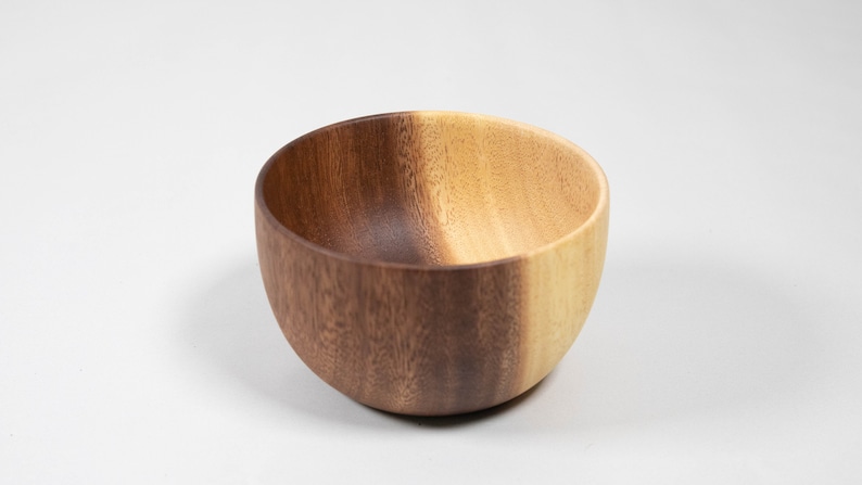 May include: A wooden bowl with a natural wood grain finish. The bowl has a rounded shape and a smooth interior. The color is a mix of light and dark brown tones. This bowl is suitable for serving food.
