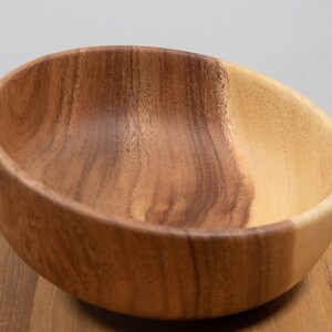 May include: A wooden bowl with a natural finish. The bowl is made of two tones of wood, with a lighter color on one side and a darker color on the other. The bowl is empty and sits on a wooden surface.