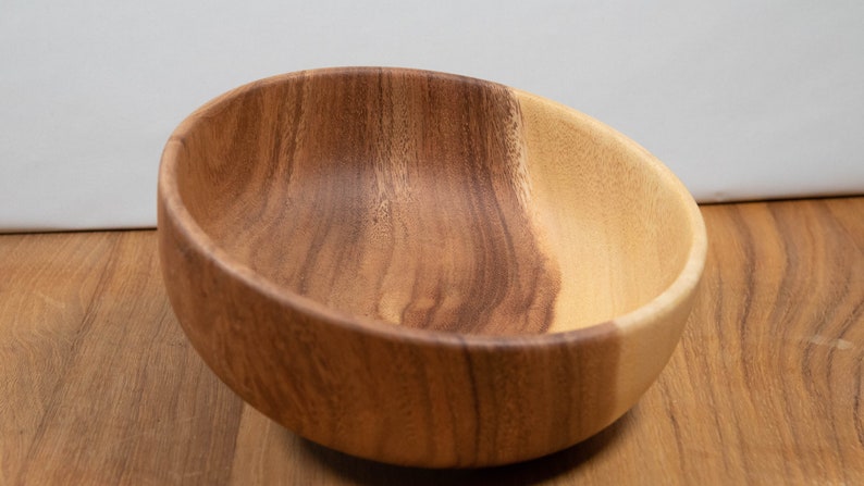 May include: A wooden bowl with a natural finish. The bowl is made of two tones of wood, with a lighter color on one side and a darker color on the other. The bowl is empty and sits on a wooden surface.