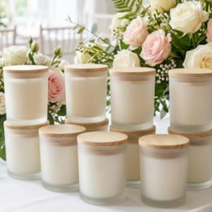 May include: A collection of ten cylindrical glass candle jars with light-colored wooden lids, each filled with white wax. The jars are arranged in a group, with some stacked slightly behind others, set against a backdrop of soft pink and white roses.
