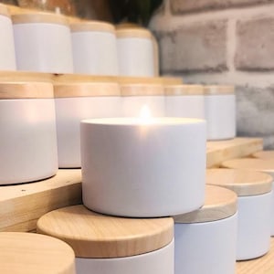 May include: A collection of white candles in various sizes, arranged on wooden shelves. The candles are in white containers with wooden lids. The candles are lit and the flames are visible.
