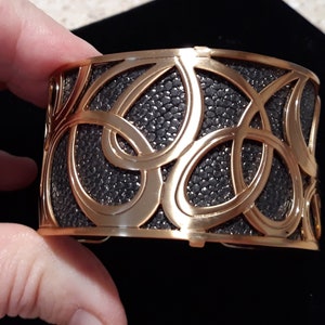 Puede incluir: Un brazalete de metal dorado con un diseño texturizado negro. El brazalete presenta un patrón intrincado de círculos superpuestos.