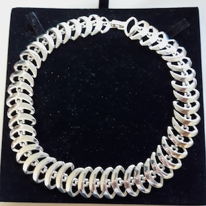 Puede incluir: Un collar de plata con un diseño único que presenta eslabones entrelazados en forma de media luna. El collar se muestra sobre un fondo de terciopelo negro.