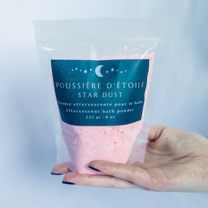 Peut inclure: Un sac en plastique transparent rempli de sels de bain roses. L'étiquette indique "POUSSIÈRE D'ÉTOILE STAR DUST" et "Poudre effervescente pour le bain Effervescent bath powder 225 gr | 8 oz".