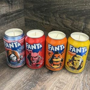 May include: Four Fanta soda can candles with horror movie character designs. The cans are red, orange, blue, and yellow. Each candle has a white wick and a silver top. The cans are on a wooden surface.