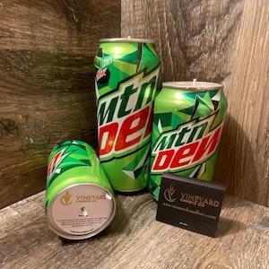 May include: Three green and white Mountain Dew-branded candles in the shape of aluminum cans. The candles are labeled "Vineyard Candle Co."