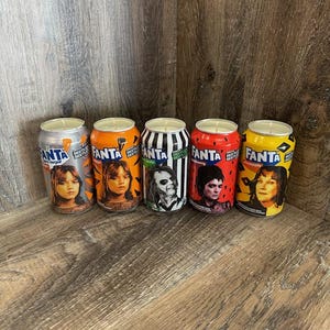 May include: Five orange and silver cans with a black and white striped can in the middle. Each can has a different character from a popular movie on the front. The cans are labeled "Fanta" and "Nestle" with the text "No Sugar" on the silver can.