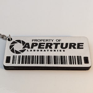 May include: A silver keychain with a black and white tag that reads "PROPERTY OF APERTURE LABORATORIES". The tag has a black and white logo of a circle with a triangle inside.