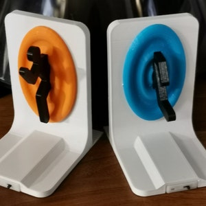 May include: A pair of white bookends with orange and blue portal designs from the video game Portal. Each bookend features a stylized human figure stepping through a portal.