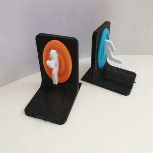 May include: Two black bookends with a 3D printed design. One bookend features a blue circle with a white figure, and the other bookend features an orange circle with a white figure.