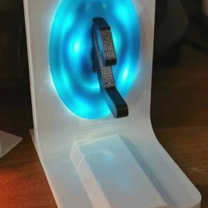 May include: A white 3D printed stand with a blue light-up circle and a black 3D printed object in the center. The stand has a flat surface on top and a curved base.